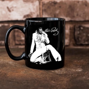 May include: Black ceramic mug featuring a white image of Elvis Presley in a performance pose. The mug has a black handle and the singer's signature in white. The background is a brick wall.