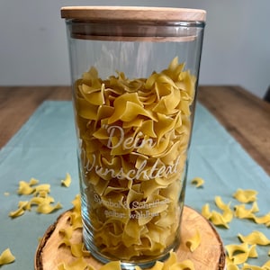May include: A clear glass jar filled with yellow egg noodles, topped with a wooden lid. The jar has etched text that reads "Dein Wunschtext" and "Symbol & Schrift selbst w&auml;hlbar". The jar sits on a wooden slice.
