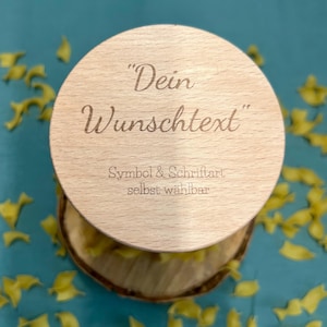 May include: A round, light-colored wooden lid with the engraved text "Dein Wunschtext" and "Symbol & Schriftart selbst w&auml;hlbar". The lid sits atop a wooden base, with a blue background and scattered yellow shapes.