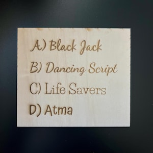 May include: A square wooden plaque with engraved text. The text includes the phrases "Black Jack", "Dancing Script", "Life Savers", and "Atma", each preceded by a letter. The wood has a light, natural color.