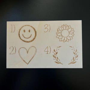 May include: A light-colored wooden plaque with four engraved designs: a smiley face, a heart, a sunflower, and a laurel wreath. Each design is numbered from 1 to 4. The designs are in a light brown color.