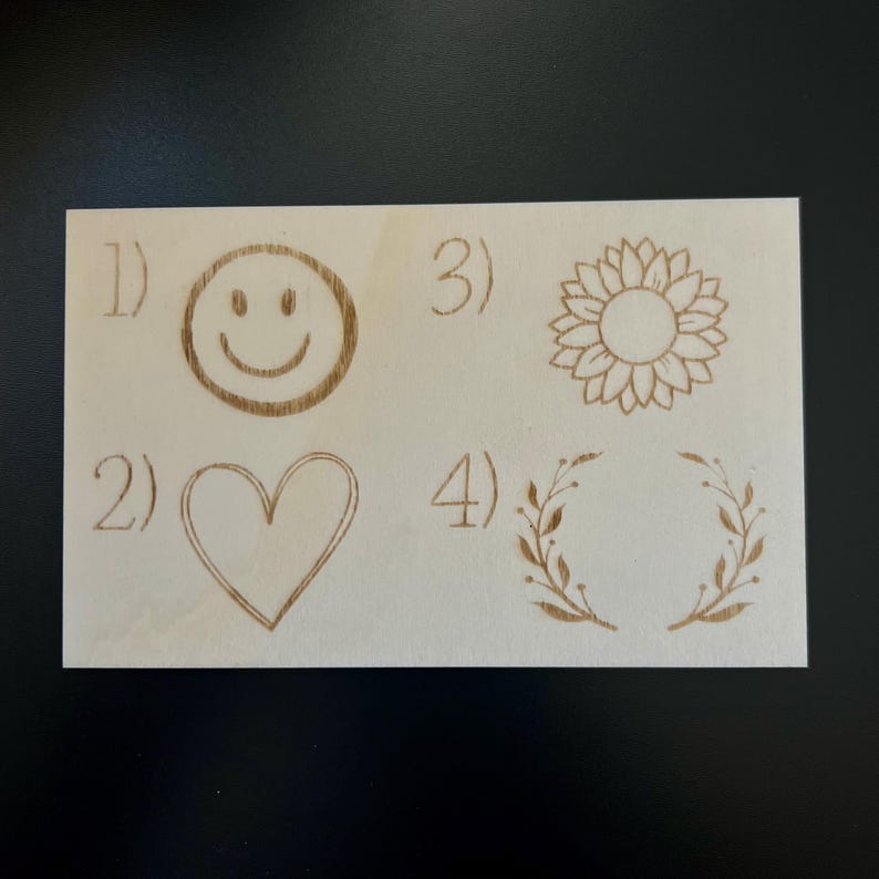 May include: A light-colored wooden plaque with four engraved designs: a smiley face, a heart, a sunflower, and a laurel wreath. Each design is numbered from 1 to 4. The designs are in a light brown color.