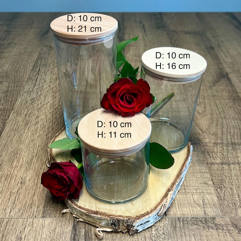 May include: Three clear glass jars with wooden lids, each 10 cm in diameter. The heights are 21 cm, 16 cm, and 11 cm. A red rose and greenery are placed near the jars. The jars are on a wooden surface.