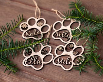 May include: Four wooden paw print ornaments with names Nala, Nelly, Ella, and Milo, each with a snowflake or flower design. The ornaments are on a brown wooden surface, with green pine branches.