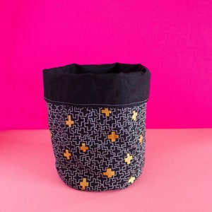 May include: Black fabric storage bin with a white and gold geometric pattern. The bin has a black fabric top and is sitting on a pink surface.