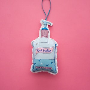 May include: A blue and white hand sanitizer bottle ornament with the text "Hand Sanitizer" and "Kills 99.9% of Germs". The ornament is hanging from a blue ribbon.