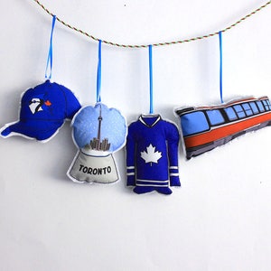 May include: Five blue and white fabric ornaments hanging from a string. The ornaments are shaped like a baseball cap, a snow globe with the Toronto skyline, a hockey jersey, a streetcar, and a penguin.
