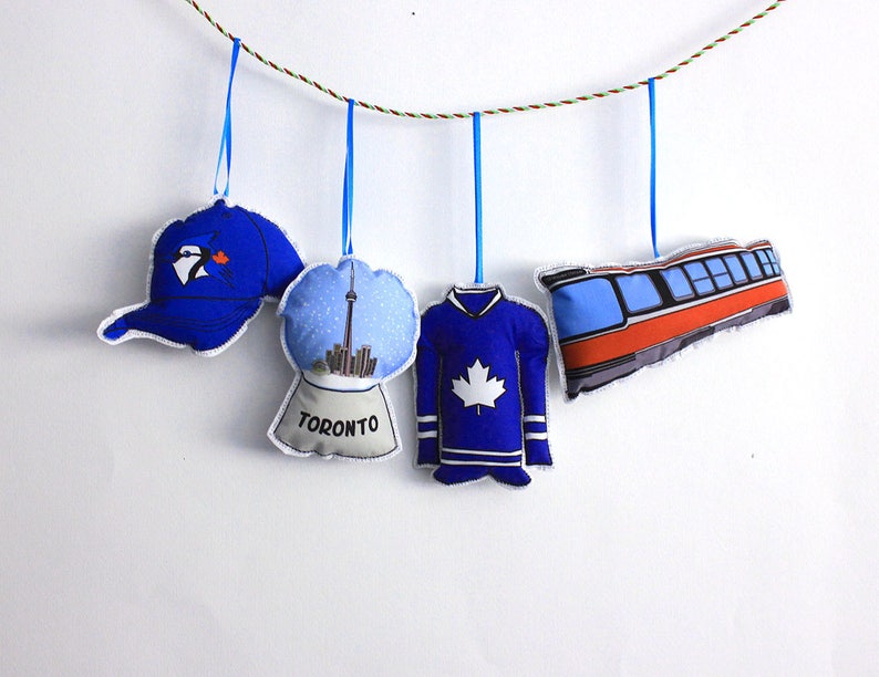 May include: Five blue and white fabric ornaments hanging from a string. The ornaments are shaped like a baseball cap, a snow globe with the Toronto skyline, a hockey jersey, a streetcar, and a penguin.