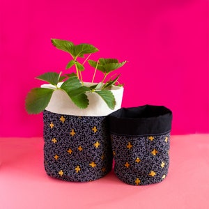 May include: Two black fabric planters with a white trim and gold plus sign pattern. The larger planter has a green plant inside.