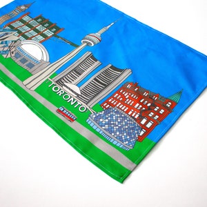 May include: A colorful illustration of the Toronto skyline on a blue and green fabric. The illustration includes the CN Tower, the Royal Ontario Museum, and other iconic buildings. The text "TORONTO" is printed in white on the green fabric.