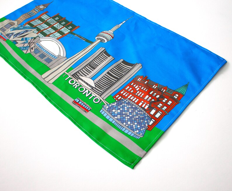 May include: A colorful illustration of the Toronto skyline on a blue and green fabric. The illustration includes the CN Tower, the Royal Ontario Museum, and other iconic buildings. The text "TORONTO" is printed in white on the green fabric.
