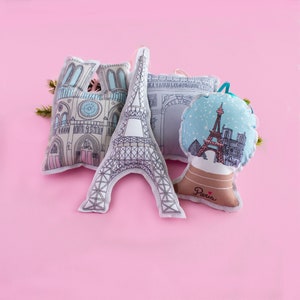 May include: Four plush ornaments featuring Parisian landmarks. The ornaments are shaped like the Eiffel Tower, Notre Dame Cathedral, the Arc de Triomphe, and a snow globe with the Eiffel Tower inside. The ornaments are all white with black outlines and have a pink background.