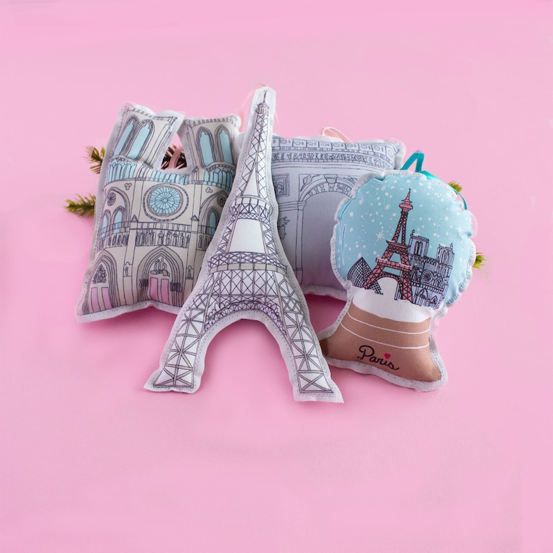 May include: Four plush ornaments featuring Parisian landmarks. The ornaments are shaped like the Eiffel Tower, Notre Dame Cathedral, the Arc de Triomphe, and a snow globe with the Eiffel Tower inside. The ornaments are all white with black outlines and have a pink background.