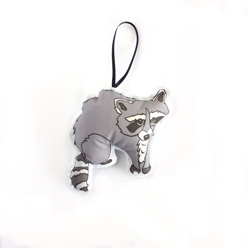 May include: A gray and white plush raccoon ornament with a black hanging loop. The raccoon has a black nose and eyes, and a white stripe on its forehead.