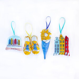 May include: Four colorful felt ornaments with different Dutch themes. The ornaments are shaped like a snow globe, a pair of clogs, a cone of fries, and a row of houses. The snow globe features a depiction of Amsterdam, the clogs have the word "Holland" on them, the fries are labeled "Dutch Fries", and the houses are labeled "Amsterdam".