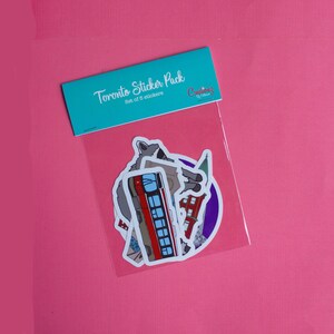 May include: A sticker pack featuring Toronto landmarks, including a streetcar, a building, and a wolf. The pack includes 5 stickers and is labeled "Toronto Sticker Pack, Set of 5 stickers, Creations by Chelsea".