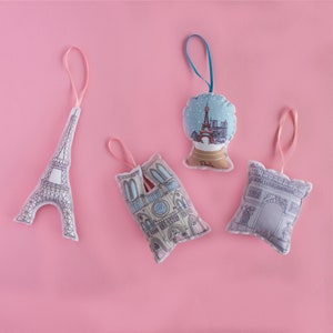 May include: Four fabric ornaments with Parisian themes. A pink Eiffel Tower, a gray Arc de Triomphe, a white Notre Dame Cathedral, and a blue snow globe with the Eiffel Tower inside. All ornaments have a loop for hanging.