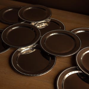 Può includere: Una collezione di piatti da portata color argento con bordo perlato. I piatti sono disposti su una superficie di legno, riflettendo la luce. I piatti sono rotondi e hanno un design semplice ed elegante, adatto per cene formali.