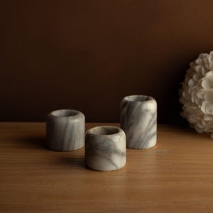 May include: Three cylindrical marble candle holders with gray veining. The candle holders are of varying heights and sit on a wooden surface. The background is a dark brown color.