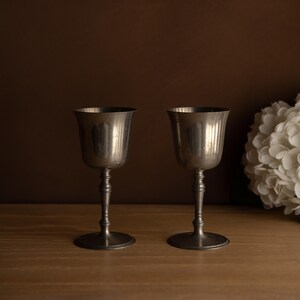 Puede incluir: Dos copas antiguas de plata con un diseño acanalado en el cuenco y un tallo detallado. Las copas se exhiben sobre una superficie de madera con un fondo marrón oscuro y un arreglo floral blanco.