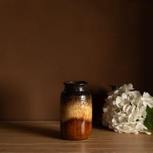 Puede incluir: Un jarrón de cerámica con un degradado de colores, que va del negro en la parte superior al beige y luego a un marrón rojizo intenso en la base. El jarrón es cilíndrico con un borde ligeramente ensanchado. Flores blancas junto al jarrón.