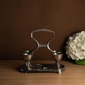 May include: A silver-toned egg holder with four egg cups and a handle. The holder sits on a round base and is displayed on a wooden surface. A white hydrangea flower is in the background.