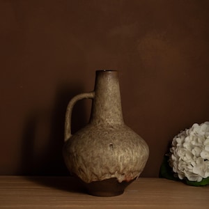 Puede incluir: Un jarrón de gres con asa, con una superficie texturizada y moteada en tonos marrones y grises. El jarrón tiene una base marrón oscuro y un cuello estrecho. Un arreglo floral blanco está a la derecha.