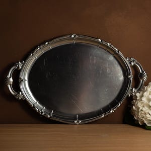 Può includere: Vassoio ovale color argento con manici decorati e bordo decorativo. Il vassoio in metallo ha una superficie leggermente graffiata. Il vassoio è su una superficie di legno con sfondo marrone.