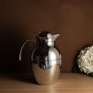 Può includere: Una caraffa da caffè argentata lucida con un manico curvo e un coperchio. La caraffa ha un corpo bulboso e un beccuccio per versare. Lo sfondo è di colore marrone scuro e sulla destra è visibile una composizione floreale bianca.