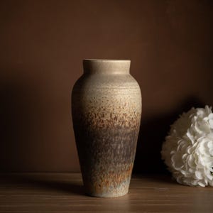 Peut inclure: Un vase en céramique de grande taille, avec une palette de couleurs terreuses et mouchetées. Le vase passe d'un beige clair en haut à un marron foncé à la base, avec des touches de vert et d'orange. Le vase est posé sur une surface en bois.