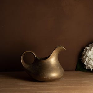 May include: A small, antique brass jug with a rounded body and a curved spout. The jug has a circular handle and sits on a wooden surface. The background is a solid brown colour, with a cluster of white flowers on the right.