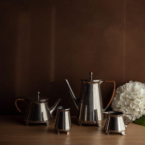 Puede incluir: Un juego de té de plata pulida, que incluye una tetera, una cafetera, una crema y un azucarero, se coloca sobre una superficie de madera. La cafetera y la tetera tienen asas tejidas. Un arreglo floral blanco está en el fondo.