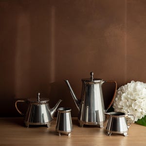 Op de afbeelding: Een zilveren theeservies met een theepot, koffiepot, melkkan en suikerpot. De set staat op een houten oppervlak tegen een bruine achtergrond. De theepot en koffiepot hebben bruine handvatten.