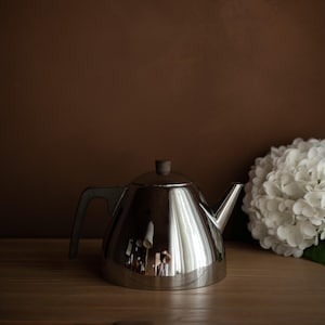 Peut inclure: Une théière en acier inoxydable brillant avec une poignée sombre et un bouton en bois. La théière est posée sur une surface en bois, reflétant l'environnement. Un arrangement floral blanc est à droite.