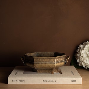 Könnte beinhalten: Eine achteckige Schale aus Messing mit zwei Griffen, die auf einem Buch mit dem Titel "bea mombaers items & interiors LUSTER" steht. Die Schale hat ein verwittertes Aussehen und liegt auf einem hellen Buch.