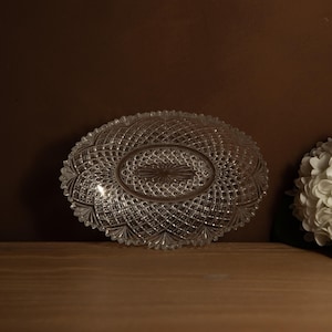 May include: An oval-shaped, clear glass serving dish with a detailed diamond pattern. The dish has a scalloped edge and a central oval design. The glass reflects light, creating a sparkling effect. The dish is on a wooden surface.