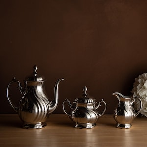 Peut inclure: Un service à thé argenté, comprenant une théière, un sucrier et une crémière, est présenté sur une surface en bois. L'ensemble présente un design cannelé et une finition polie. Le fond est marron foncé.