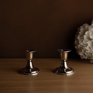 Peut inclure: Deux bougeoirs argentés au design classique. Chaque bougeoir a une base large et ronde avec un bord perlé, une tige fine et un petit dessus circulaire. Les bougeoirs sont présentés sur une surface en bois.