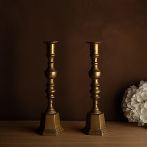 Peut inclure: Deux bougeoirs anciens en laiton. Chaque bougeoir présente une base hexagonale, une tige détaillée et un sommet en forme de coupe. Les bougeoirs sont exposés sur une surface en bois devant un fond marron foncé.
