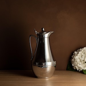May include: A polished silver pitcher with a curved handle and a spout, set against a brown backdrop. The pitcher has a lid and a bulbous base. A white hydrangea flower is on the right side of the image.