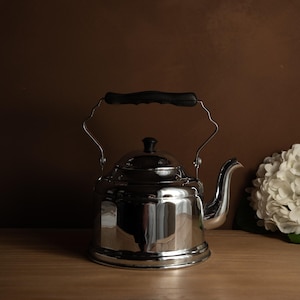 Może przedstawiać: Błyszczący czajnik ze stali nierdzewnej z czarnym uchwytem i gałką. Czajnik ma zakrzywioną wylewkę i stoi na drewnianej powierzchni. Biała kompozycja kwiatowa znajduje się w tle.