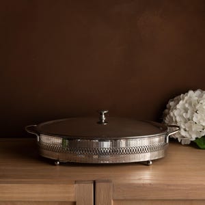 Könnte beinhalten: Eine silberfarbene, ovale Servierschale mit Deckel. Die Schale hat ein dekoratives, durchbrochenes Design am Boden und kleine Füße. Der Deckel hat einen kleinen Griff. Die Schale steht auf einer Holzoberfläche.