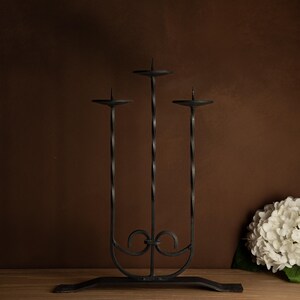 May include: A black wrought iron candelabra with three candle holders. The candelabra has a decorative base and twisted stems. The candle holders are circular and have a spike in the center. The candelabra is approximately 12 inches tall.