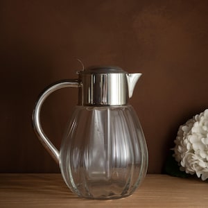 May include: A clear glass jug with a silver metal lid, handle, and spout. The jug features vertical ridges and is placed on a wooden surface. A white flower arrangement is in the background against a brown wall.