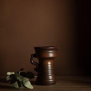 Può includere: Una brocca in ceramica marrone scuro con manico e beccuccio. La brocca ha una superficie strutturata ed è posta su uno sfondo marrone. Un rametto di foglie verdi poggia sulla superficie di legno davanti alla brocca.