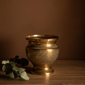 May include: A decorative brass vase with detailed floral engravings. The vase features a wide rim and a pedestal base. The ornate carvings cover the body of the vase. It is positioned on a wooden surface. A sprig of green leaves is in the foreground.