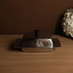Puede incluir: Una mantequera de acero inoxidable con base rectangular y tapa a juego. La tapa tiene un pomo negro en la parte superior para levantarla fácilmente. El plato está sobre una superficie de madera.