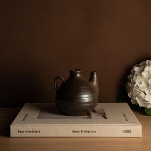 Może przedstawiać: Ciemnobrązowy ceramiczny dzbanek z uchwytem i dziobkiem, umieszczony na książce zatytułowanej "items & interiors" autorstwa bea mombaers. Dzbanek jest ustawiony na ciemnobrązowym tle, z białym układem kwiatowym po prawej stronie.