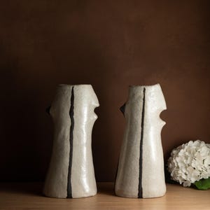 Peut inclure: Deux vases en céramique blanc cassé avec une rayure verticale noire et un design unique en forme de visage. Chaque vase a une base évasée et une surface texturée. Les vases sont exposés sur une surface en bois sur un fond marron.