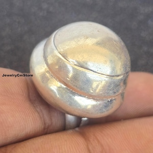 May include: A silver ring with a distinctive design is shown in close-up. The ring has a rounded top and a band that encircles the finger. The ring is made of a shiny, metallic material.